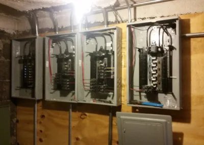 Three open electrical panels showing wiring and circuit breakers on a basement wall.