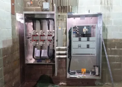 Industrial electrical panels with open doors revealing wiring and components, installed against a concrete block wall.