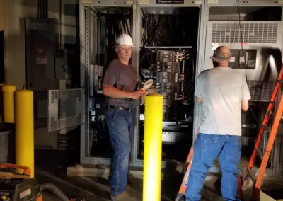Two electricians working on electrical panels in an industrial environment.