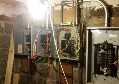Three electrical panels with exposed wiring installed on a basement wall with visible signs of dampness and mold.