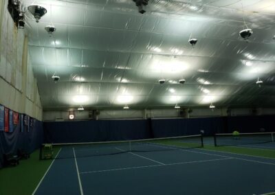 An indoor tennis court with multiple overhead lights, a net in the center, and a basket of tennis balls on the left side. The ceiling is covered with reflective material.