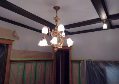 A vintage chandelier with ornate design hangs from a ceiling with dark beams. The room is under renovation, with plastic sheeting covering walls and tape around the edges.