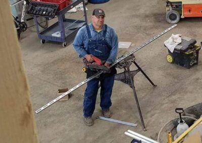 A person wearing overalls and a cap is working with a metal bar on a sawhorse stand in a workshop. Various tools, barrels, and equipment are visible around the workspace.
