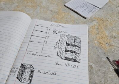 A hand holding a notebook open to a page with hand-drawn sketches and measurements of a furniture piece. A folded piece of paper and a screwdriver are on the sawdust-covered table.