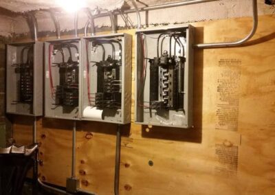 Three open electrical panels mounted on a wooden board, with visible circuit breaker components and wiring. The room has unfinished walls and a ceiling light.
