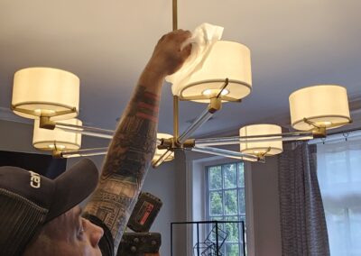 Person wearing a cap and with visible tattoos on their right arm cleans a modern chandelier using a cloth. A ladder is partially visible in the background near a window.
